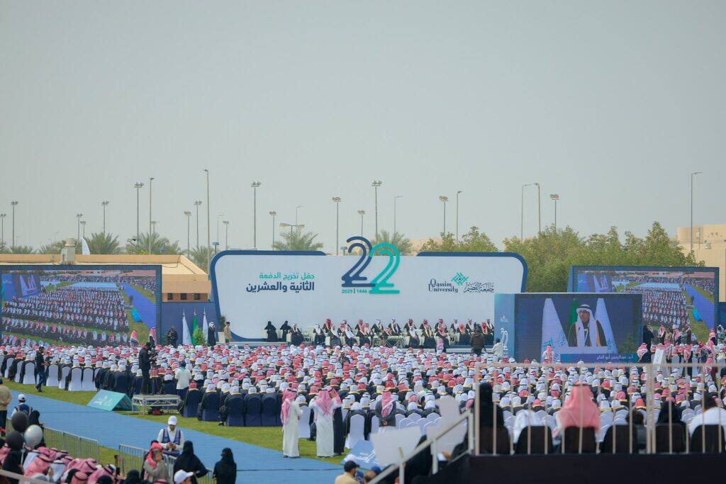 A large outdoor graduation for the graduates, with a large number of participants dressed in white clothes with the red and white Saudi Shemagh or Ghutra, heading towards the stage of the "22" production and an Arabic performance. The ceremony was attended by His Highness the Emir of Qassim Province.