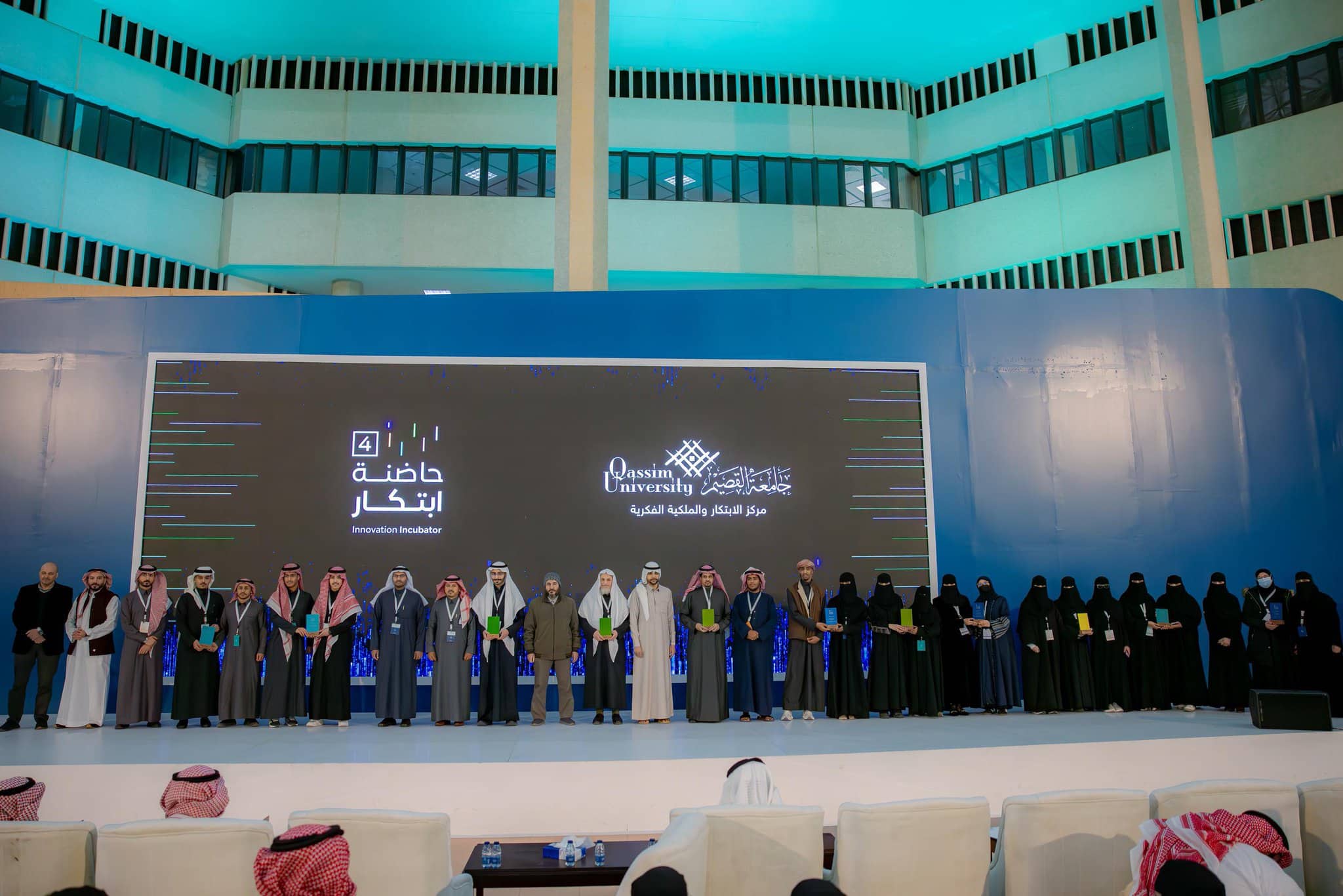 A large gathering of men and women in traditional Saudi attire, with the men wearing the shemagh or Saudi ghutra, gathered at the Innovation Incubator Hackathon's award podium. Behind them is a large screen with the Qassim University logo and prominent text in Arabic highlighting the participating projects.