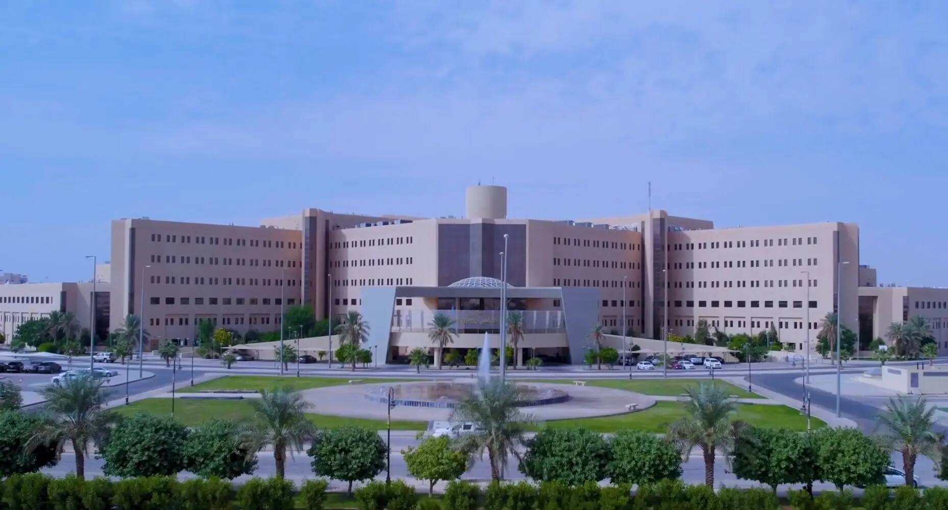 A luxurious and modern government building, consisting of several floors, stands tall amidst carefully landscaped green spaces and rows of palm trees. The building features a spacious central entrance, adorned with a roundabout and an elegant fountain, reflecting the values and ambitions of Saudi Vision 2030. Set under a clear blue sky, visitors and faculty members are dressed in traditional Saudi attire, including the shemagh or ghutra, emphasizing the institution's commitment to national identity and excellence, befitting the stature of Qassim University.