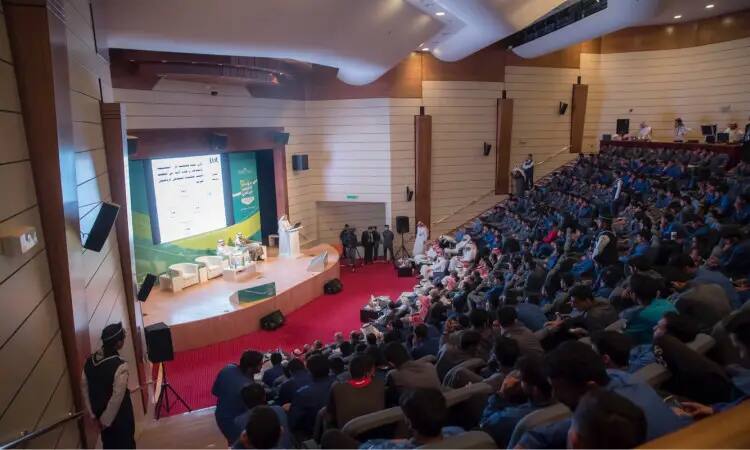 A large crowd prepares to gather in the Al-Qassim Lecture Hall, their main attention focused on the stage, where a large screen shows a presentation on the topic of sports fanaticism. Many of the attendees are dressed in formal Saudi attire, sporting the kabara or Saudi shemagh with the Saudi aqal and bisht. The auditorium's lighting and modern design is characterized by wood paneling and luxurious red carpets.