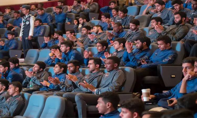 A large group of young men in matching blue jackets, wearing the Saudi Arabian ghutra or shemagh with an aqal, and some in Saudi winter, sit in a lecture hall in Qassim. The audience watches with interest the abstract events unfolding in the international competition, applauding in appreciation of what is presented in front of them, in a scene that expresses the Kingdom's spiritual Thai sport in a focused and disciplined manner.