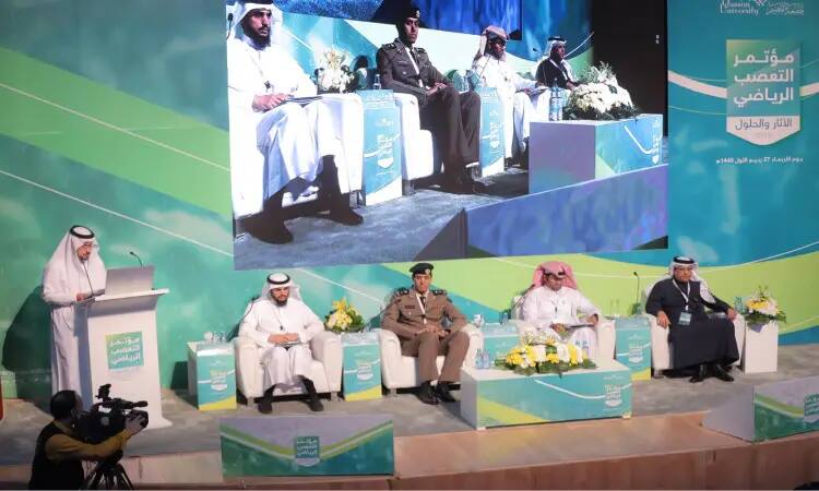 Five men, wearing the Ghutra or Shemagh and Eqbal, and the Saudi Bisht, were seated on a podium at an event at Qassim University. One of them walks up to the podium to face the officials, while the image of the committee is projected on the big screen behind them. In the background, banners in Arabic and English with slogans discussing "Sports in the Kingdom," coordinated in a decorative medium dominated by the color green, effectively conveys a sense of official responsibility.