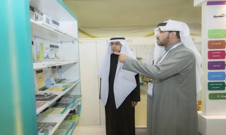 A man wearing a Saudi Ghutra or Shemagh with a Saudi Eqal and Bisht inside a pavilion at the Qassim Book Fair looks at the books and pamphlets on the shelves. He points to the term, as he continues to point to the right panels.