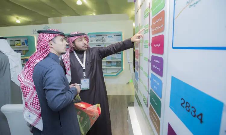 Two men dressed in Saudi Arabian tunic, headband and bisht examine an information board at the Qassim Book Fair, where details about the contents of a bookstore are indicated by colorful diagrams and text in Arabic.