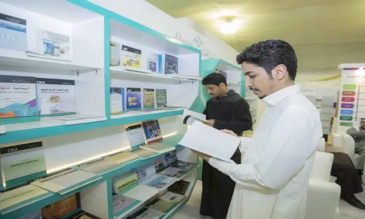 Wearing the Ghutrah and Shemagh, the official Saudi bisht, in front of the bookshelves at the Qassim Book Fair in Qassim. All of you are engrossed in reading books, respecting the contemporary culture of the library and highlighting the diversity of its cultural contents, including a focus on science and knowledge.