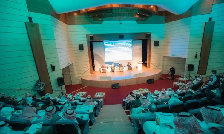 A symposium or panel discussion, such as an international conference on developing the performance of a university professor, is held in a lecture hall with a predominantly male audience wearing the Saudi dress with the Saudi ghutra or shemagh topped with the Saudi bisht. Attendees go face-to-face and a large screen displays texts and materials related to the topic of the discussion.