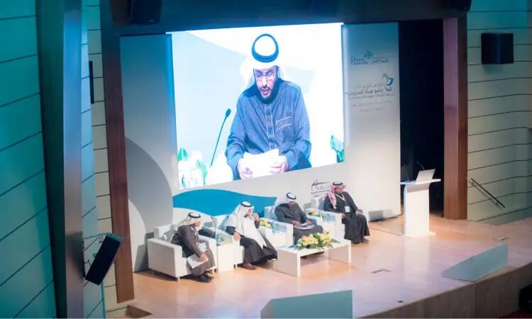 Four men in a Saudi Ghutra or Shemagh with a Saudi Eqal and Bisht sit on the podium during conference sessions, while someone delivers a speech from the pulpit. On the big screen behind them should be a picture of a documentary speaker during the International Conference for the Evaluation of Faculty Performance at Qassim University.