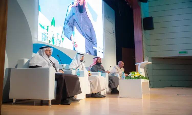 The four men under the Saudi ghutra, aqal and bisht sit on the stage of the international conference at Qassim Hall, discussing the development of the university professor, with flasks in front of them. Behind them is a large screen with a related topic, suggesting a conference or the image of a children's scientific seminar.