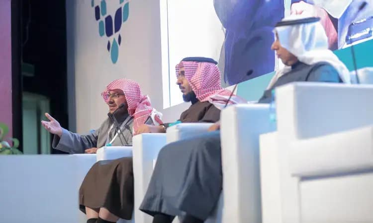 يجلس ثلاثة رجال ينتظرون الغترة أو الشماغ والعقال والبشتاء السعودي على منصة حمراء بكراسٍ بيضاء، حيث ينخرطون في نقاش رسمي ضمن مؤتمرات مؤتمرات نحو مجتمع إيجابي في القصيم. يظهر أحد واضحين وهو يشير بيده أثناء حديثه، بينما يستمع للآخرين باهتمام. وتبدو خلفهم شاشة كبيرة وتصميمات تجريدية بارزة في خلفية الحدث.