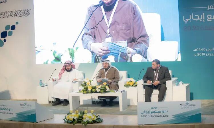 Three men sit at the podium, wearing the Saudi tunic or shemagh with the Saudi headband and bisht, with microphones and papers in hand during the scientific sessions of the Vision 2030 conference at Qassim University. Behind them, the main communication network takes center stage, while the blue and white color scheme and floral arrangements are scientific, highlighting the values of the community.