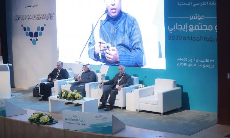 Men sit on stage during a community entertainment conference, with a large board behind them bearing Arabic texts and the logo of the Kingdom's Vision 2030. One man steps up to the podium wearing a Saudi bisht and a ghutra or shemagh with an aqal, while white chairs in the auditorium adorn the tables with bouquets of flowers, as part of events in Qassim.