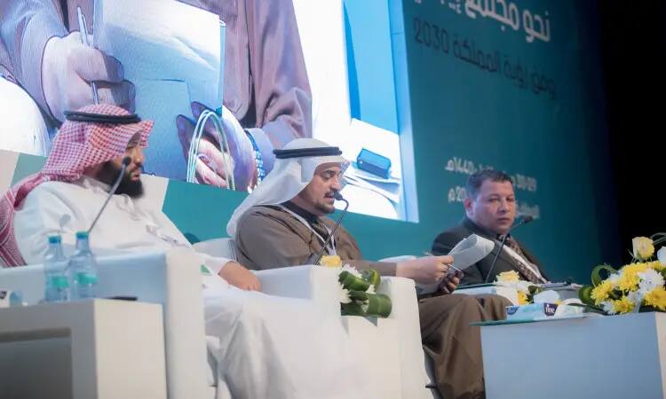 Three men sit on stage during the Vision 2030 conference in Qassim, two of them wearing the Saudi Ghutra, Eqal and Bisht, while the third is wearing a men's suit. In front of them, they are adorned with the smell of wine and glass. Behind them on the big screen are the details of the scientific sessions written in Arabic.
