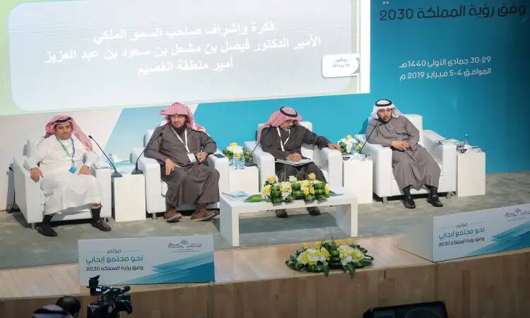 Surrounded by white chairs and fresh flowers, four men in Saudi bishtar, ghitra or shemagh and aqal sit during the Vision 2030 conference at Qassim University. Behind them are banners with phrases such as "Positive Society" and "Scientific Sessions," as well as the Vision 2030 logo displayed as an invitation to the conference.