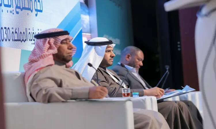 Three men sit at the podium, dressed in traditional Saudi attire in the Ghutra or Saudi Shemagh, with the aqal and the official Saudi bisht. In front of each of them are papers and cups of tea. The banner bears Arabic text referring to the event as part of the Children's Vision 2030 conference. The banner focuses on scientific sessions and promotes the concept of a leadership community, as part of the official organization in Qassim.