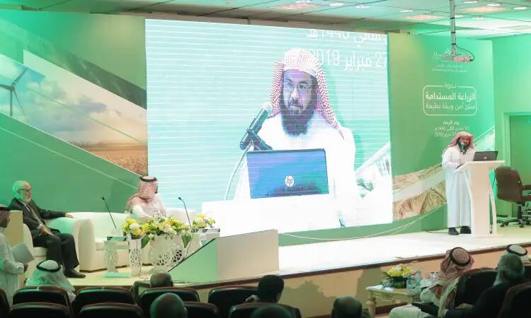 One of them, dressed in a Saudi ghutra, aqal, and bisht, addresses the audience from the stage, using a laptop, while his image is clearly projected on a large screen behind him. A group of designers sit at the tisse table, leaving the backdrop with images of wind turbines and text in Arabic close to the themes of sustainability in agricultural milk.