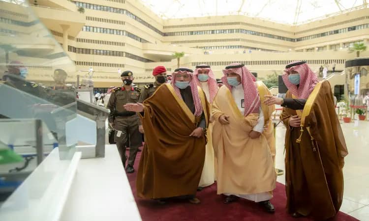 A group of men wearing the Saudi shemagh or Saudi ghitra and headband, as well as the Saudi bisht, gather in a spacious lobby in Qassim. They are chatting and watching something with interest, while behind them are security personnel, observing the formalities.