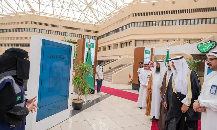 A number of men dressed in Saudi Arabian ghutra or shemagh, aqal and bisht, along with a woman in a niqab, call into an internal hall at Qassim University. A large digital screen is set up in the space, and Saudi Arabia's flags and flag symbols are prominently and proudly displayed under a raised dome out of respect and national pride.