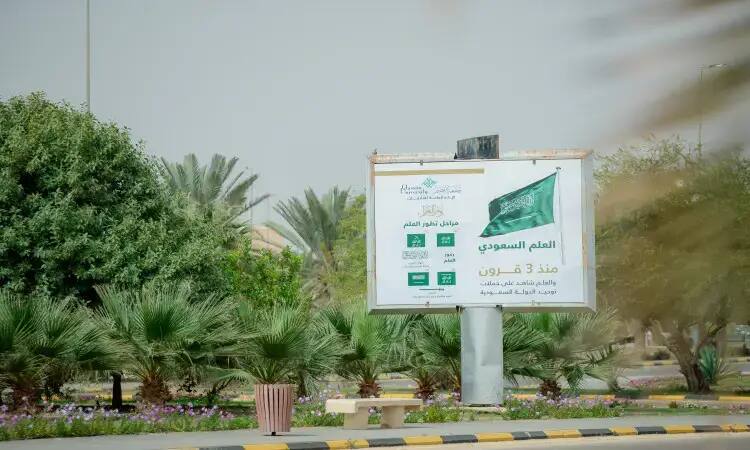 In an official scene in Qassim, a billboard stands in the middle of an area landscaped with four plants and palm trees, displaying the flag of the Kingdom of Saudi Arabia with Arabic phrases under the flag's day. Standing next to the billboard are a number of eight men dressed in the elegantly embroidered Saudi bisht, wearing on their heads the shemagh or ghutra and aqal in an image that epitomizes national authenticity. Complemented by the presence of the recognized black and white stripes.