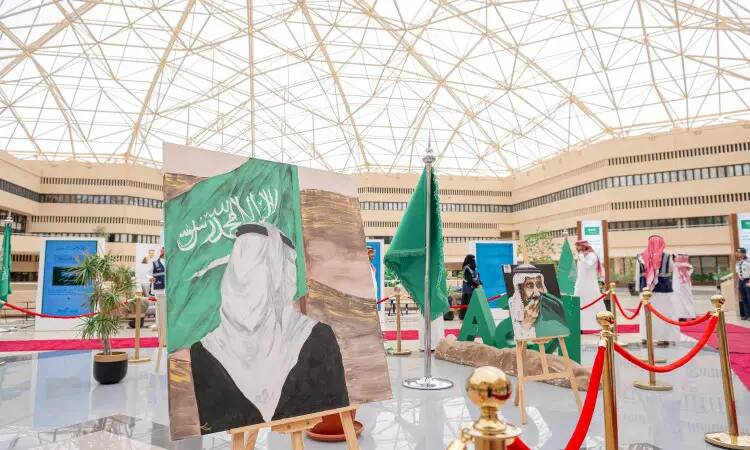 A modern pavilion specially prepared for exhibitions at Qassim University features paintings of Saudi symbols, green Saudi Arabian flags, and plaques highlighting the significance of Flag Day. Within an area delineated by red velvet barriers, visitors walk around wearing the Saudi Shemagh or Saudi Ghutrah, with the Saudi Eqal and Bisht organized; the work is designed to express pride in national identity.