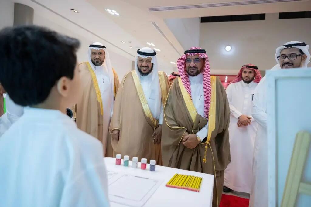 A group of men dressed in Saudi Arabia's Ghutrah, Shamaa, Aqal and Bisht, smiling and watching intently as one of the others from the event, a young boy, made contact with a screen displaying a bottle and pencils, during an event held at the seventh summer club in Al-Qassim.