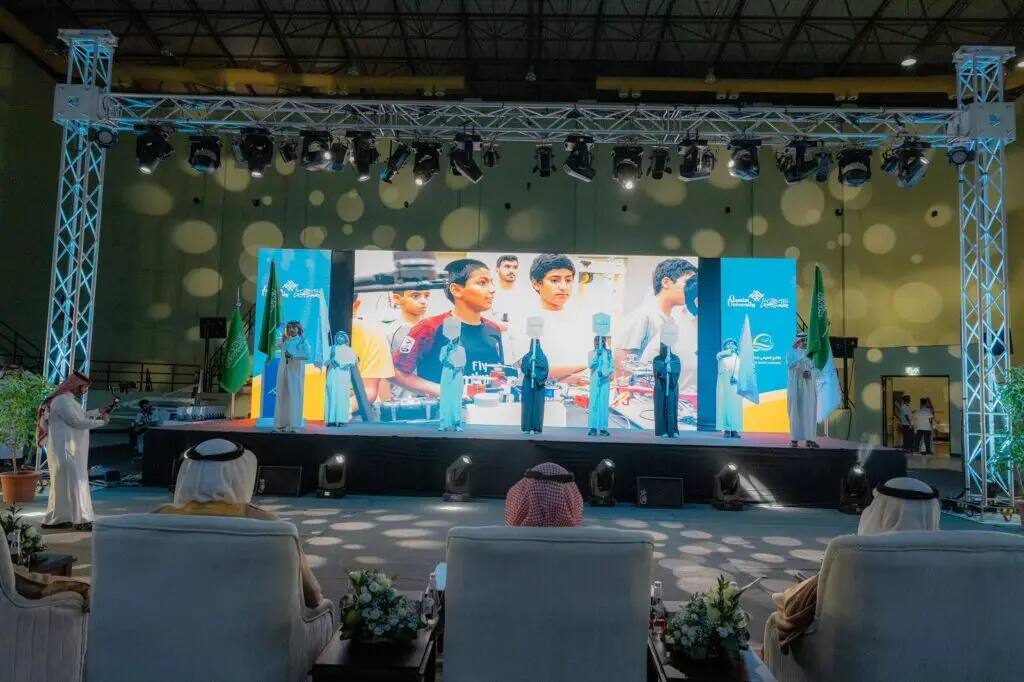 A group of people in traditional attire, including the Saudi Arabian Ghutra or Shemagh, Egal and Pasht, sit facing the stage at Qassim University, where projectors and large screens showcase the efforts of those involved in a robotics project during the seventh summer club. Bright lights illuminate the scene in a majestic and solemn scene.