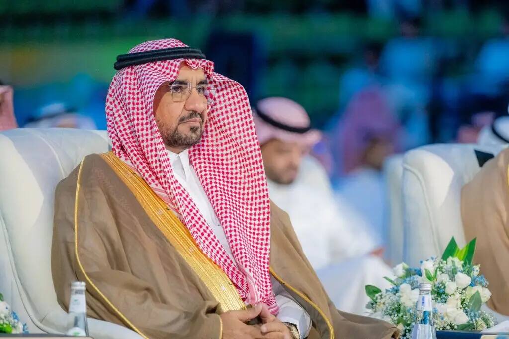 A man wearing the Saudi bisht, and wearing a white Saudi Ghutra with a red shemagh and an aqal, sits in an event organized by Qassim University as part of the activities of the seventh summer club, surrounded by a number of attendees and behind them in an irregular format in the background.