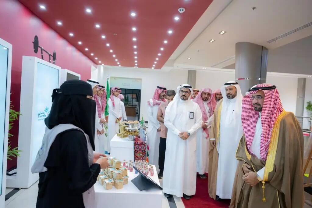 A group of men in a Saudi Arabian tunic or shemagh with an aqal and bisht, officials of the seventh summer club at Qassim University, watch as a girl presents small programs on a display table inside a brightly lit and elegantly designed enclosed exhibition hall.