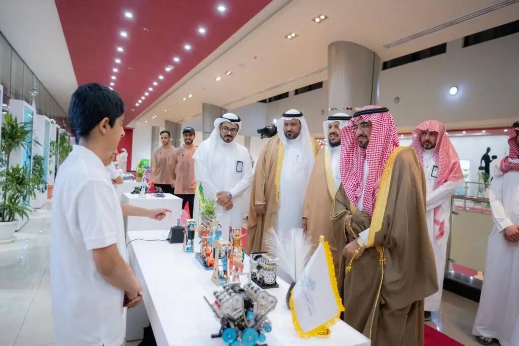 A number of men in their distinctive native dress, a Saudi tunic or shemagh with a Saudi aqal and pasht, gather at Qassim University, where they watch intently as an audience member demonstrates robotic inventions on a table inside a plush conference room. Some of the attendees are smiling and interacting with the presentation.