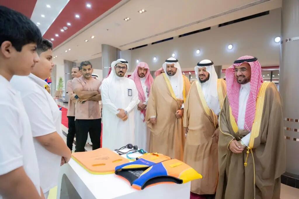 A group of men dressed in Saudi Arabian Ghutra or Shemagh with Saudi Egal and Bisht, gathered around a table displaying car accessories, interacting with a number of children as white-shirted violinists, during an event held in one of the indoor pavilions as part of the 7th Summer Club activities when Al Qassim.