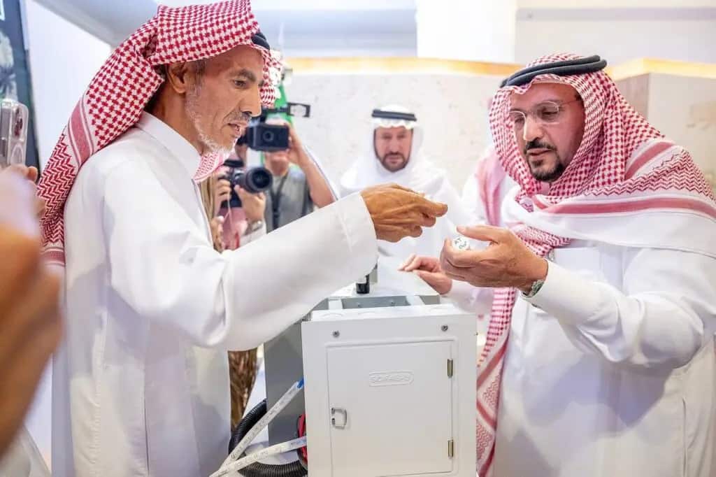 Two men dressed in Saudi Arabian ghutra, aqal and beshtet are examining an exhibit inside a university gallery, while another person calls a photographer and takes pictures in the background. A device on display in the exhibition hall is lowered between the two main subjects.