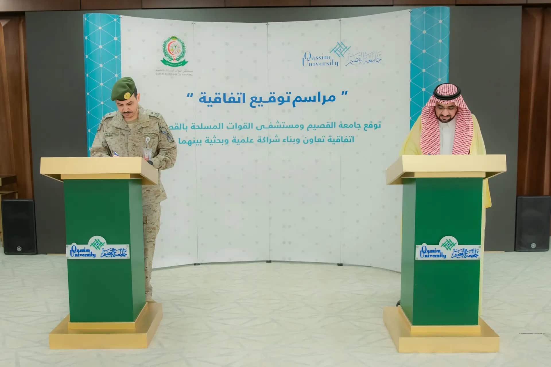 Kat is a military officer alongside a man dressed in the Saudi bisht, ghutra and aqal, all at an official podium, where they are signing a customized signature for cooperation. In the background is a painting with Arabic texts and the logos of the Armed Forces Hospital, as well as the official logo of Qassim University.