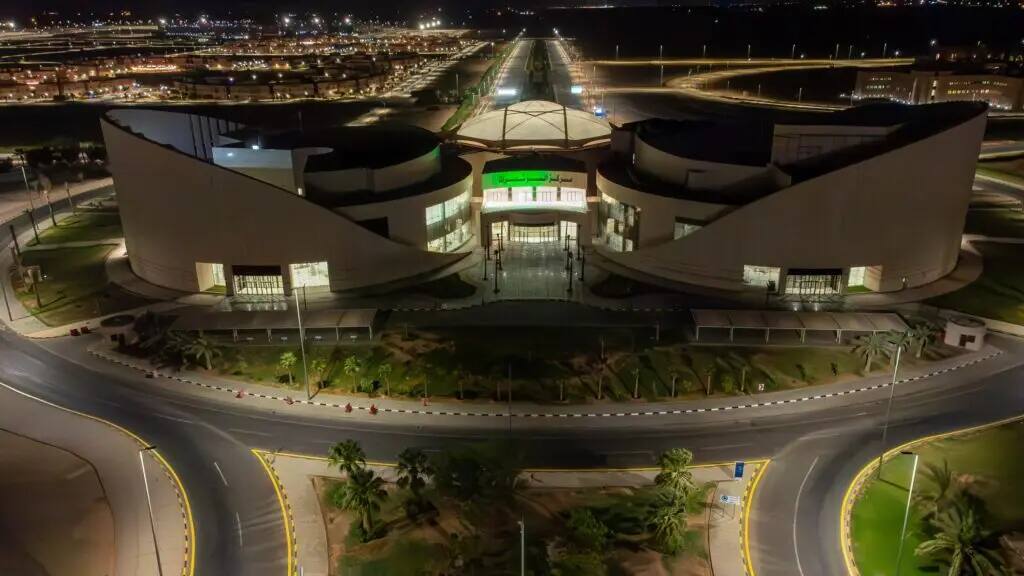 A nighttime aerial view of Qassim University's modern and elaborately lit building, a unique curved architectural design, complete with carefully landscaped roads and green spaces. The lights of the city shimmer in the background during the National Day events, where attendees appear in their official Saudi attire of Ghattara or Shemagh with Eqal and Saudi Bisht, reflecting the national identity.
