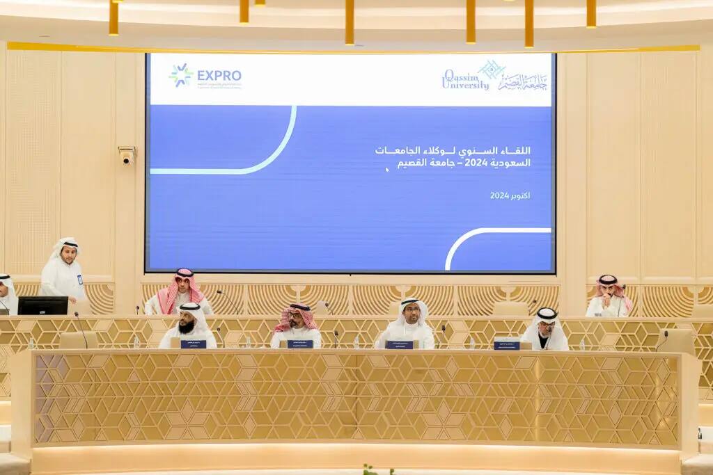 Six men in Saudi jitters, shemaghs, headbands, and pashot sit around a conference table with microphones, while a celebrity meets at the podium. A large screen broadcasts Arabic text and the slogans "Qassim University" and "Saudi Proxies at the Meeting."