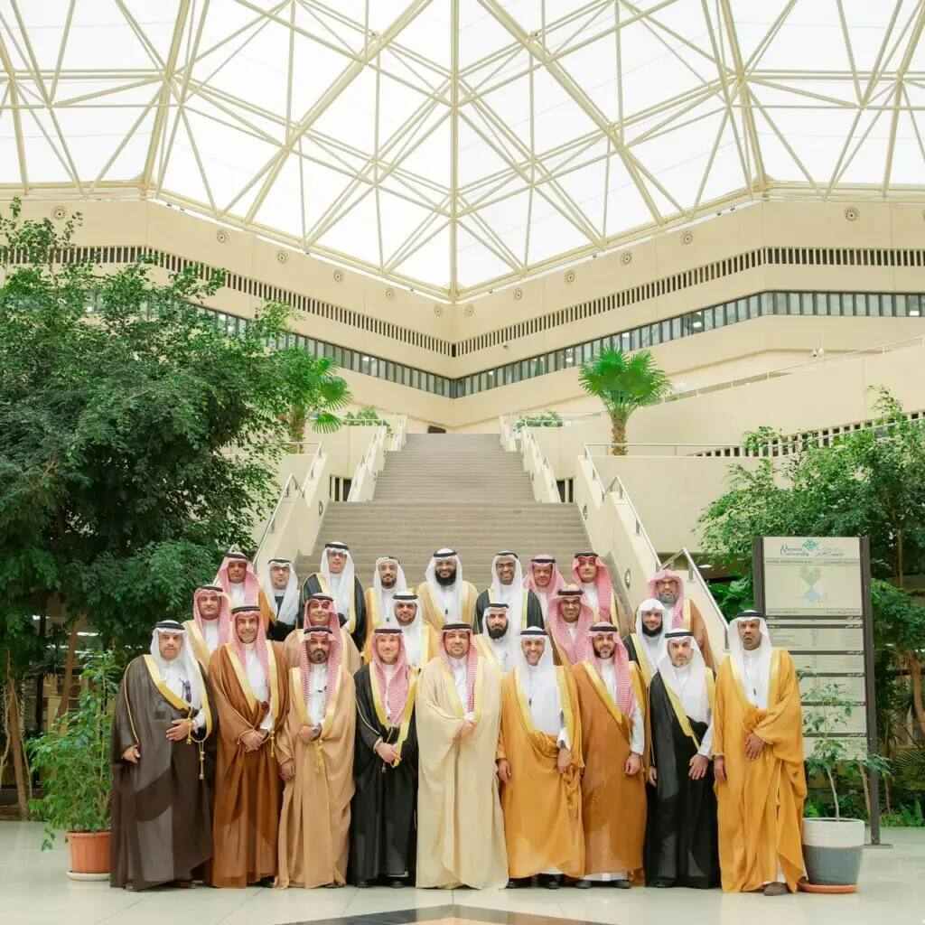 A group of men wearing the Saudi Ghutra or Shemagh with Eqal, and some wearing the official Saudi Bisht, gathered in one of the halls of Qassim University. The gathering was part of a meeting, with attendees standing in front of the staircase and indoor plants with green plants and crowning the place with a commercial glass cup, in the university's national and official cultural center.