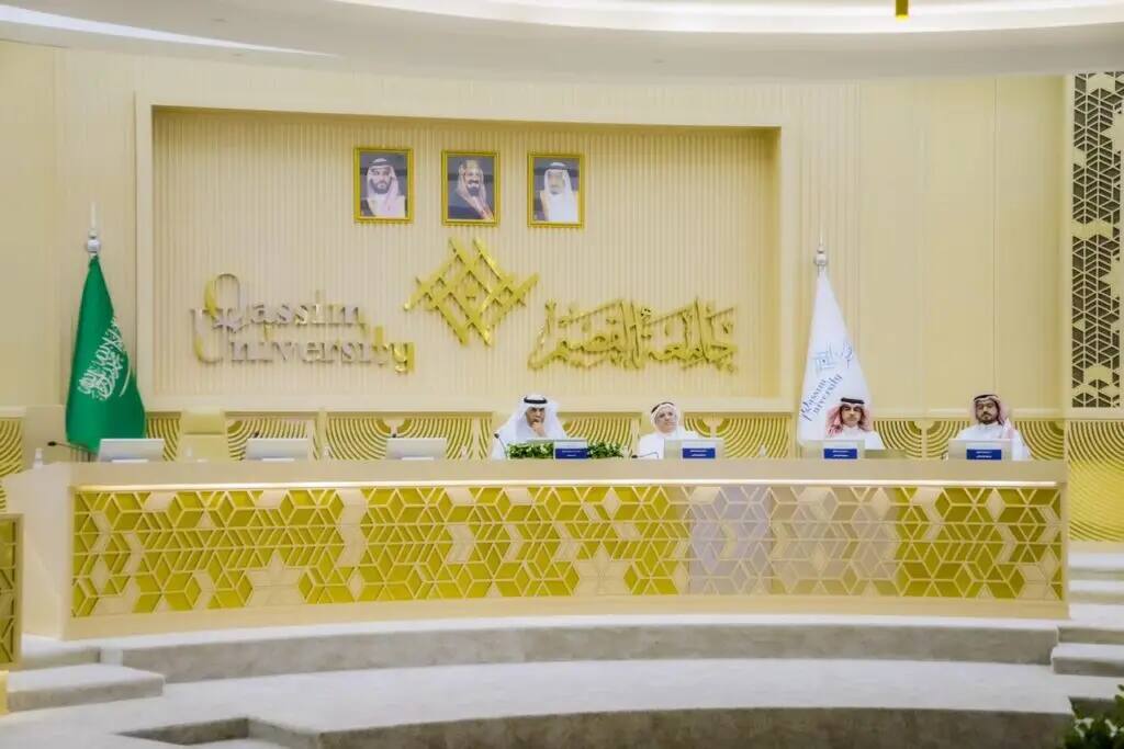 The grand conference hall at Qassim University is populated by agents of Saudi universities, where three men in shemagh or ghutra and aqal, as well as the Saudi bisht, sit behind a gold engraving stand. Behind them are the flags of Saudi Arabia and Qassim University, and a wall is lined with images of the kingdom's ruler.