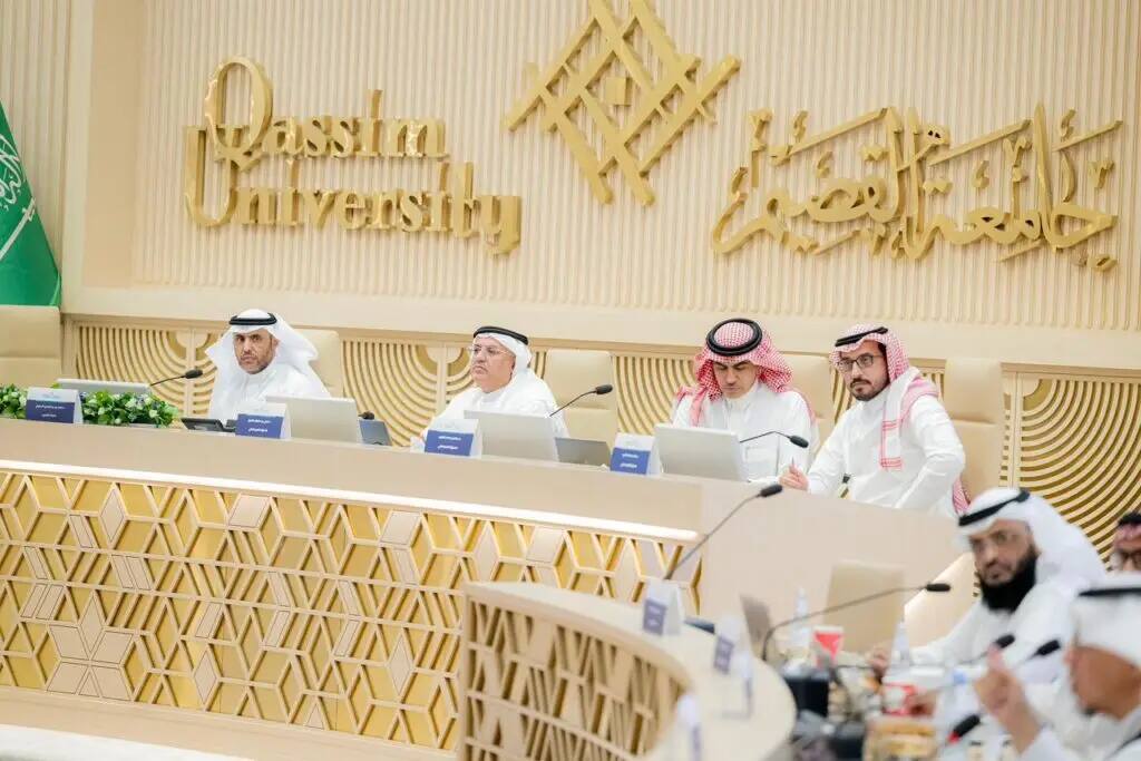 A group of men dressed in Saudi Arabian ghutra or shemagh, aqal and bisht around a long dining table eating with computers, while attending a meeting at Qassim University. The flags of the Kingdom of Saudi Arabia and the name of the university in Arabic and English are registered behind them.