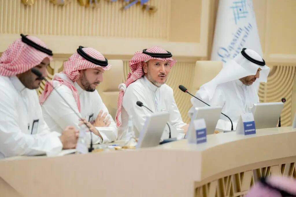 Men dressed in Saudi Arabian ghutra or shemagh with aqal and Saudi bashta sit around four conference tables with microphones and computers, participating in a computer discussion session as part of a house meeting. The Qassim University logo appears in the background, emphasizing the presence of Saudi agents and academic importance.