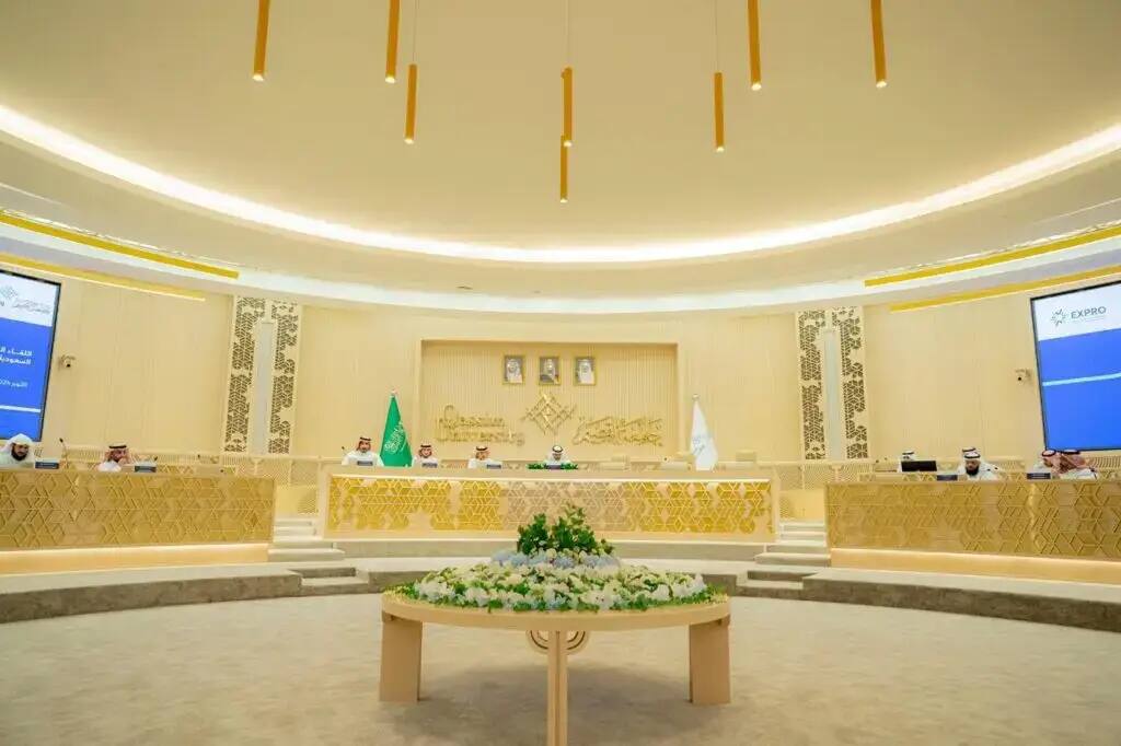 The meeting takes place in the evening in a spacious conference room, with attendees seated around a long, curved wooden table, dressed in the Ghutra or Shemagh and Eqbal with the Saudi Bisht, and the ensuing ambiance. Moments behind them, two banners protrude as screens display the EXPO logo. The hall is adorned with its official Qassim University image, and the quartet is adorned with a snowboarder.