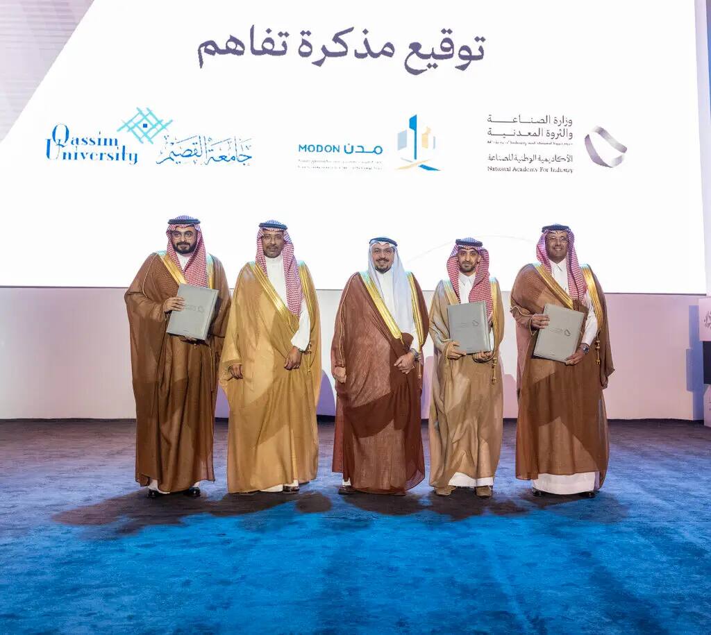 Five men wearing the Saudi ghutra or shemagh with an aqal, and some wearing the Saudi bisht, pose for a group photo in front of a background bearing the logos of Qassim University, the Saudi Industrial Cities Authority, the National Academy of Industry, and a number of other teams.