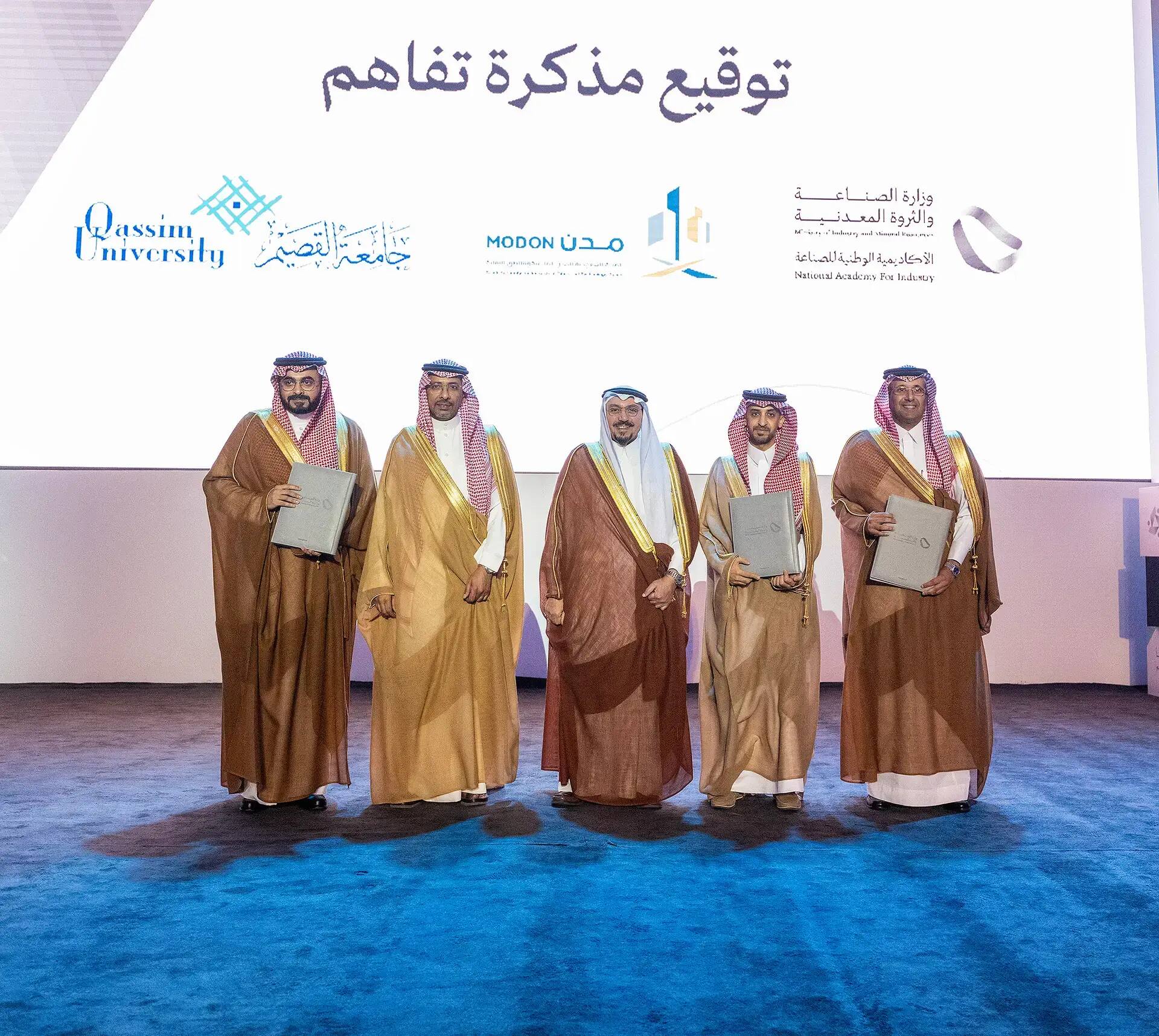 القطة خمسة رجال مرتدين الغترة أو الشماغ السعودي مع العقال، وبعضهم يرتدي البشت السعودي، على تيست حاملين وثائقي رجالي، ليلتقطوا صورة جماعية أمام خلفية تحمل شعارات جامعة القصيم والهيئة السعودية للمدن الصناعية والأكاديمية الوطنية للصناعة وعدد الفرق من الأخرى.