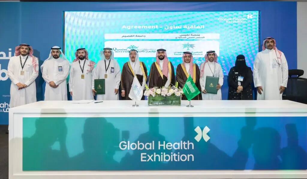 A group of people in formal attire, originating from the Saudi Shemagh or Ghutra, connect behind a table on documents and flags during the World Health Expo. This remarkable scene stands out in the exchange of experiences in medical studies and research, as a group photo is taken in front of a blue background with logos and texts in Arabic and English.