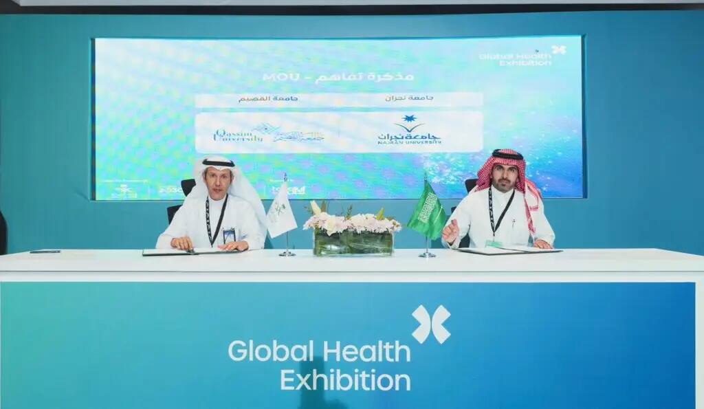 Two men in traditional Saudi attire, wearing the Saudi Shemagh, sit at an accounting table with microphones where they sign documents documenting medical studies from the World Health Trials. A screen displayed the logos of Qassim University and Majmaa University.