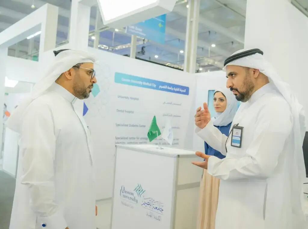 Three famous old Saudi Arabian men, from 15 Ghutrah, Shamakh, Aqal and Bashta, chatting at the event booth of the Medical Academy at Qassim University. One of the men is shown explaining with his hand during the conversation, while the other man listens intently to the woman, standing nearby. The pavilion's platform was utilized for medical information in both Arabic and English.