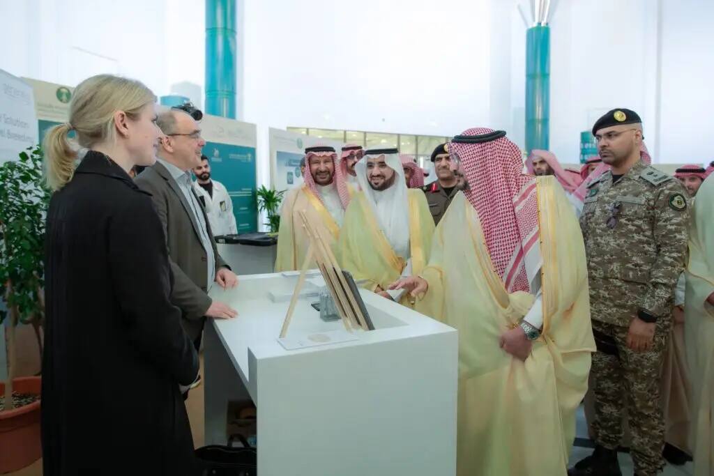 تواجد مجموعة من الرجال الحاضرين الزي السعودي التقليدي في الشماغ أو الغترة، إلى جانب أحد الجنود، حيث كانوا يتحاورون مع شخصين في منصة داخل قاعة المعرض، وذلك خلال الفعاليات العلمية الدولية الثانية للإيبل. في خلفية الشاشة عدد من الملصقات التعريفية والنباتات، مما يعكس الطابع التنظيمي والمحتوى العلمي الذي حصل على النتيجة تحت إشراف جامعة القصيم.