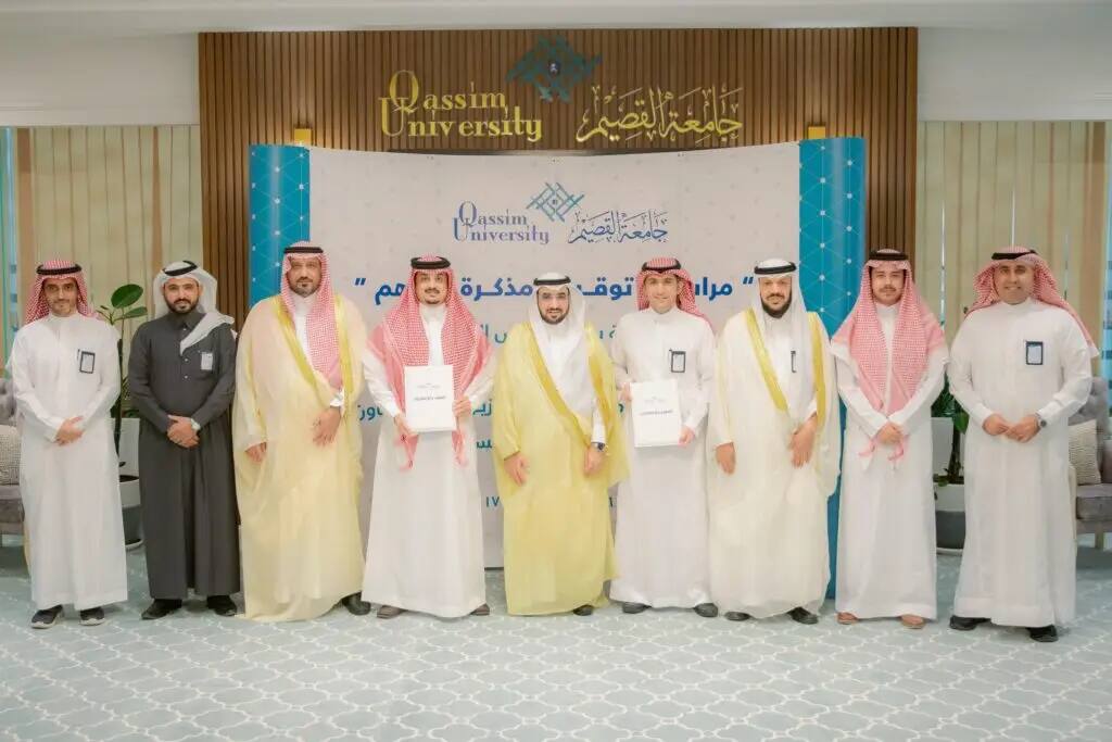 يصطف عدد من الرجال مرتدين الشماغ أو الغترة السعودية والأزياء الرسمية بالإضافة إلى البدل في مكان مغلق، وصورة جماعية أمامك وتحمل شعار جامعة القصيم ولوحة كلية الأعمال والاقتصاد. تحضر في وسط الصورة مرتين من الحضور بدقيقة الفهم.