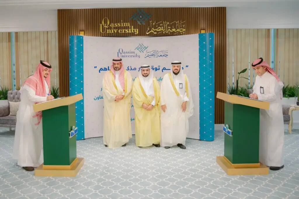خمسة رجال تولي الزي السعودي الأصلي، بما في ذلك الشماغ أو الغترة السعودية، الاتصالون في القاعة بينما يقوم اثنان منهم بتوقيع مذكرة عند تفاهمات المنصات. ملحوظة خلفهم شعار جامعة القصيم، مما يبرز اشتراكا بين شركة إقرار كلية الأعمال الشاملة في جميع الأعمار ومهنية.