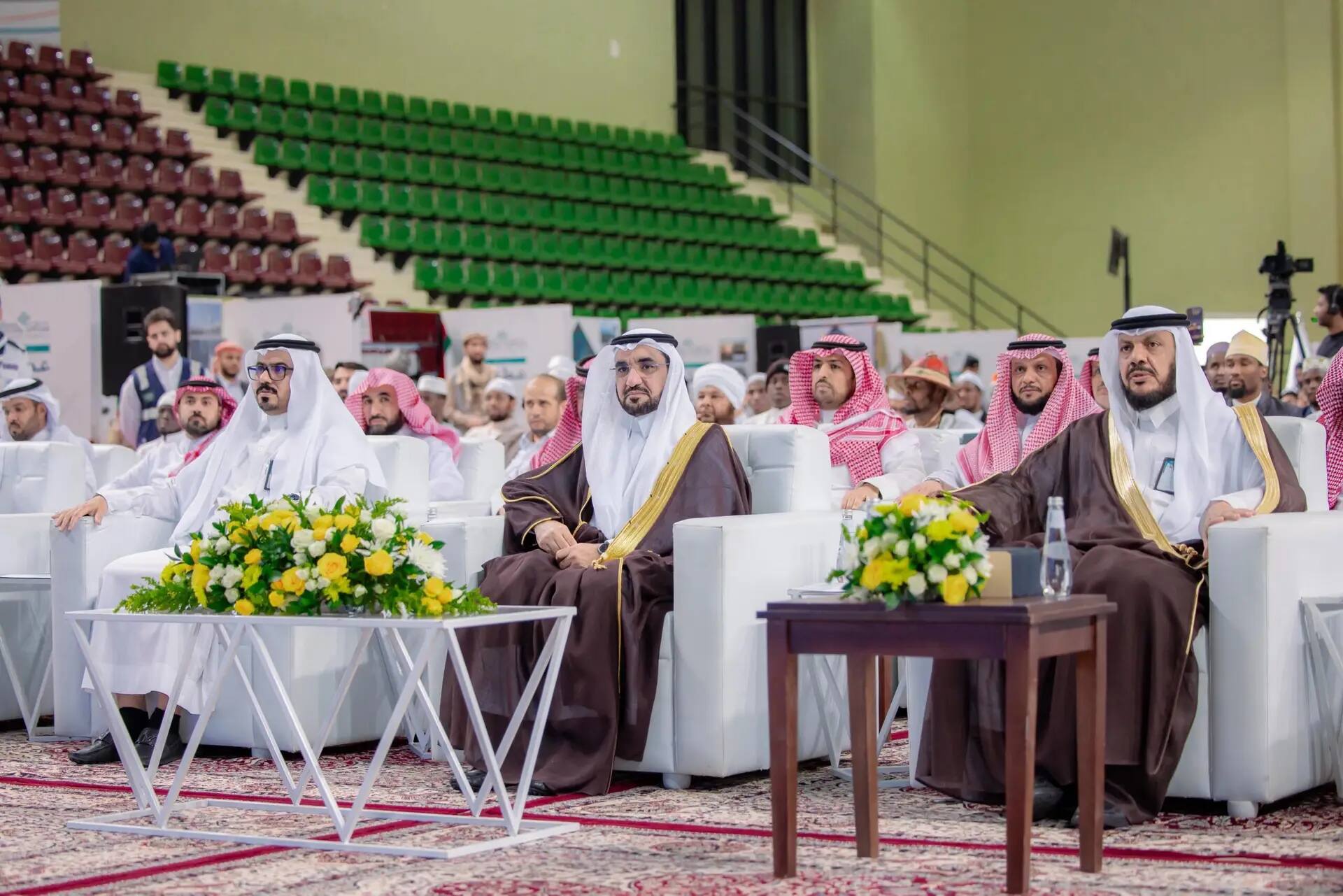 يجتمع عدد من رجال مرتدين الزي السعودي الرسمي، بما في ذلك الشماغ أو الغترة السعودية، على كراسٍ ثلاثة خلال فعالية عطاء في القصيم. وتزين الجداول تنسيقات الزهور، بينما تعيش في الخلفية المدرجة بقاعدة خضراء وحمراء.