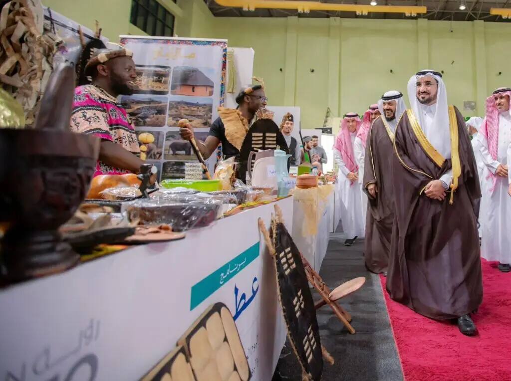 مجموعة من الرجال تولى الزيّ السعودي الأصلي، بما في ذلك الشماغ أو الغترة السعودية، يمرون بجوار جناح مخصص لتخطيط الثقافة الإفريقية والمأكولات اليدوية والأطعمة ضمن فعالية داخلية أُقيمت للاختيارة في جامعة القصيم. ويتميز موظفو الجناح باهتمامهم بزاهية الألوان وتفاعلهم مع الزائرين.