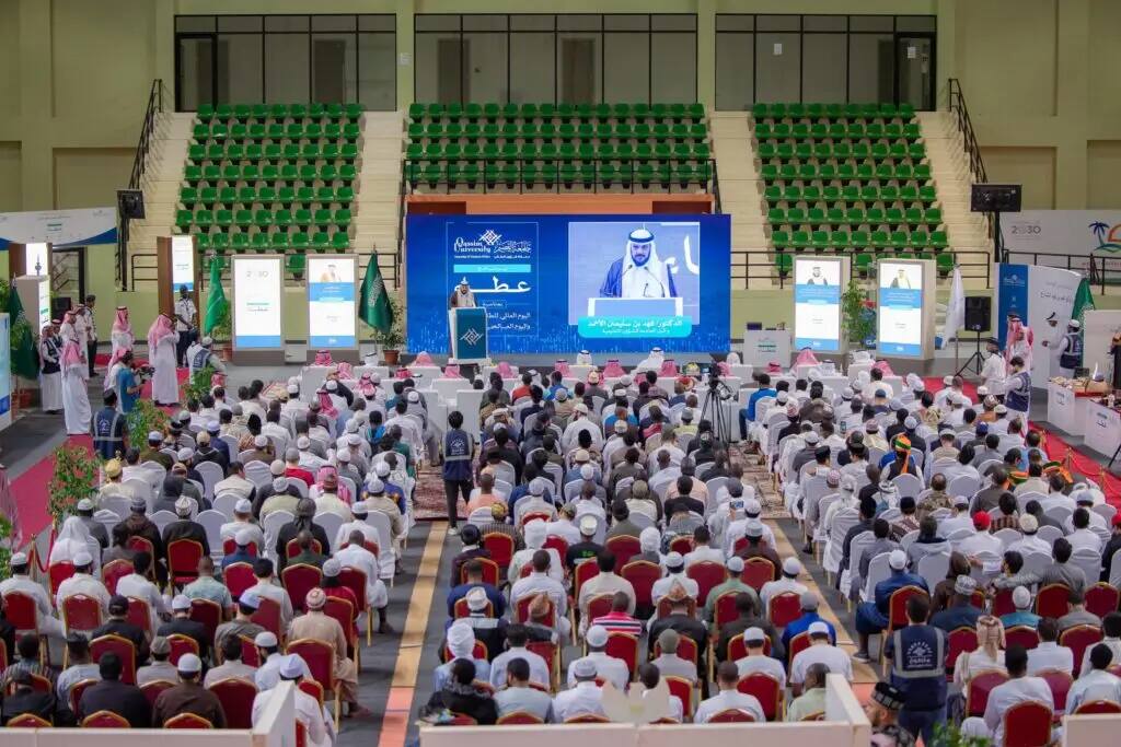 تشهد القاعة الداخلية حضورًا كثيفًا من المشاركين، يجلسون في صفوف منظمة تواجه المنصة الرئيسية، حيث تلمس معالي رئيس جامعة القصيم خلف منصة الإلقاء. وعرضت صورته بشكل ملحوظ على شاشة كبيرة خلفه. كونيتيكت للمقعد الاخضر وتوزع فيها نصوص لافتة للانتباه باللغة العربية.