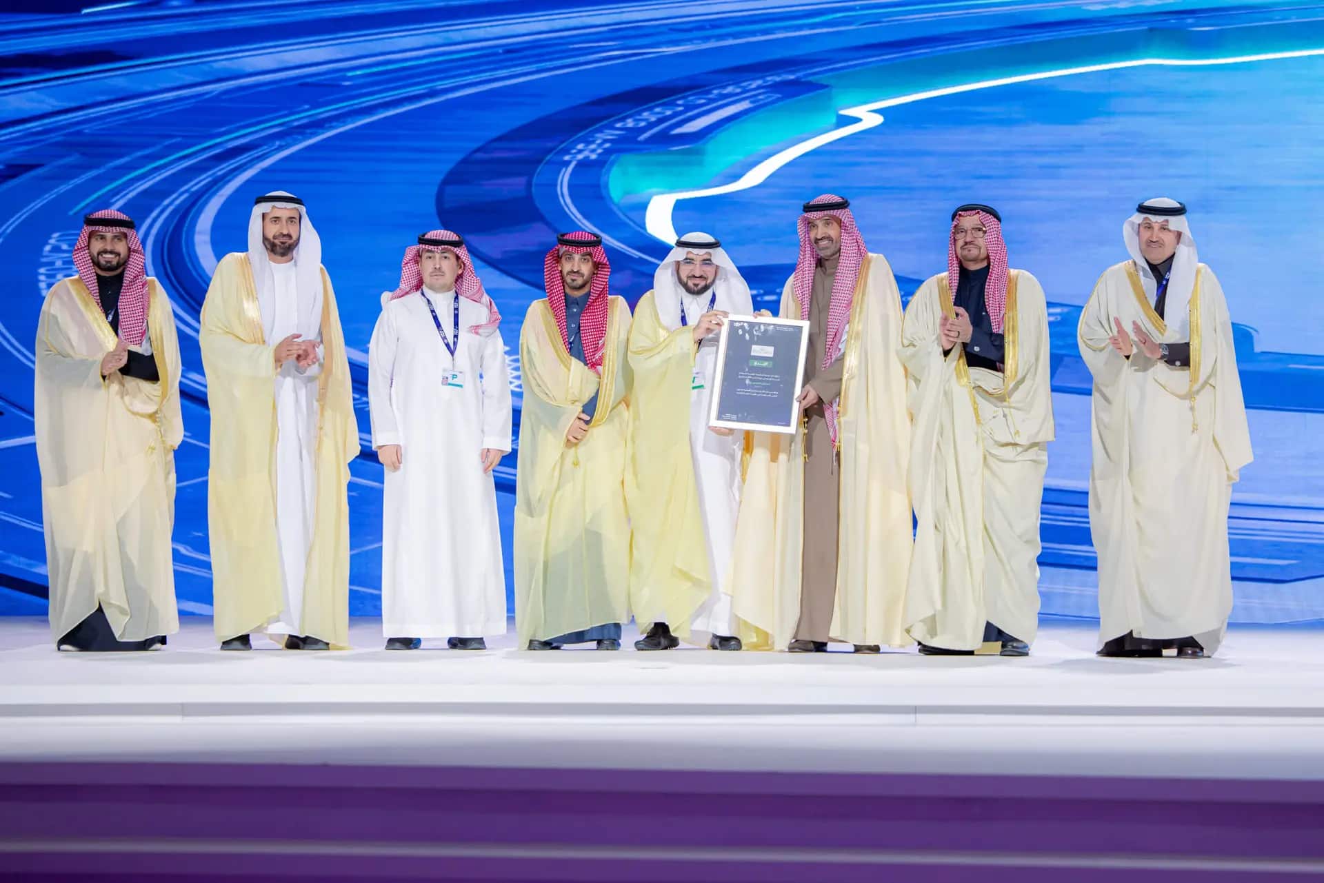 A number of men wearing Saudi shemaghs or ghattaras as well as suits of armor stood on the stage in front of a blue digital backdrop of Qassim University's digital transformation.